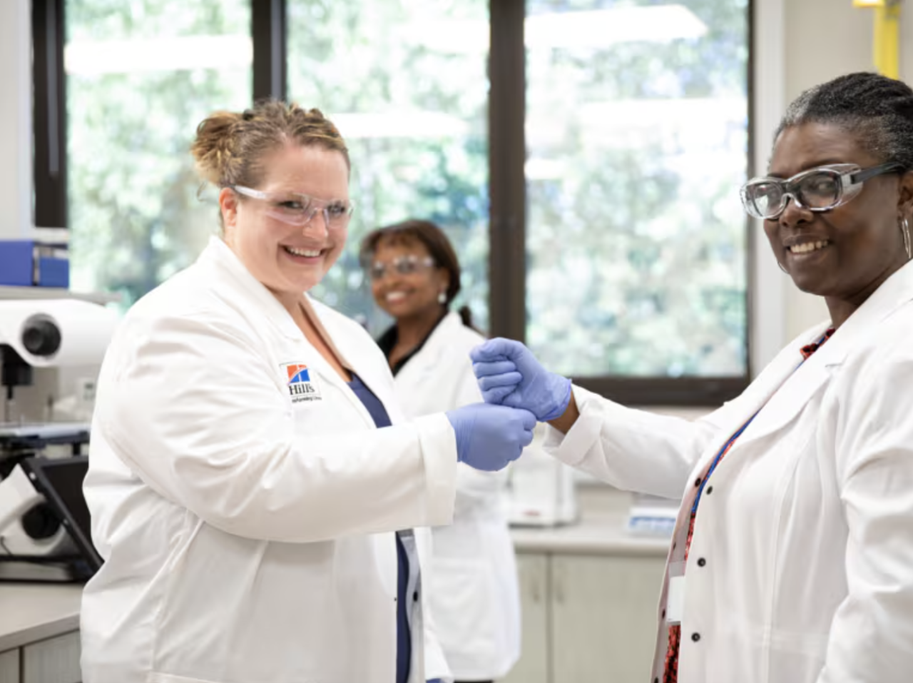 three smiling women, scientists in white coats and protective glasses